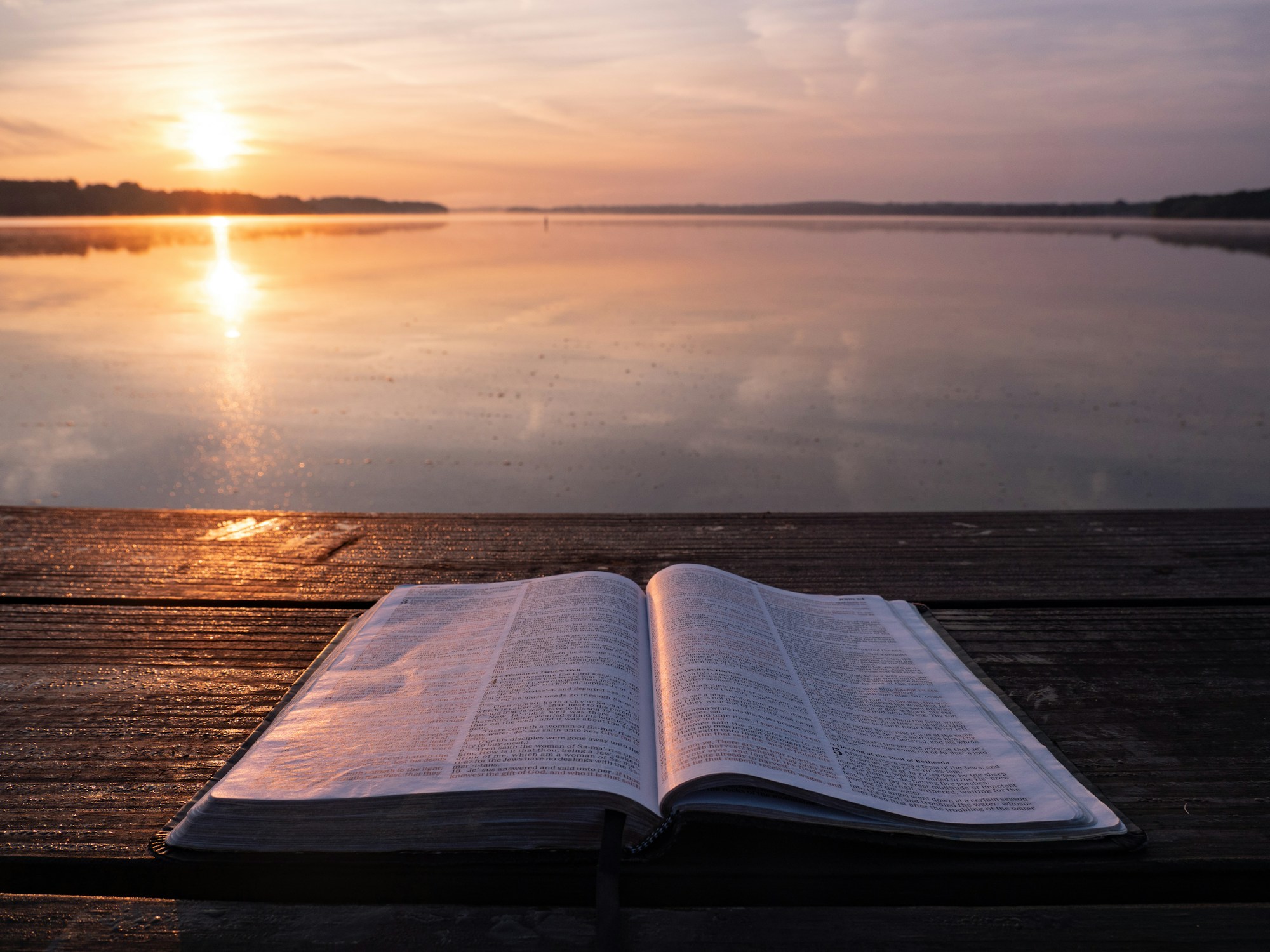Open Bible on a wooden dock at sunrise over calm water
