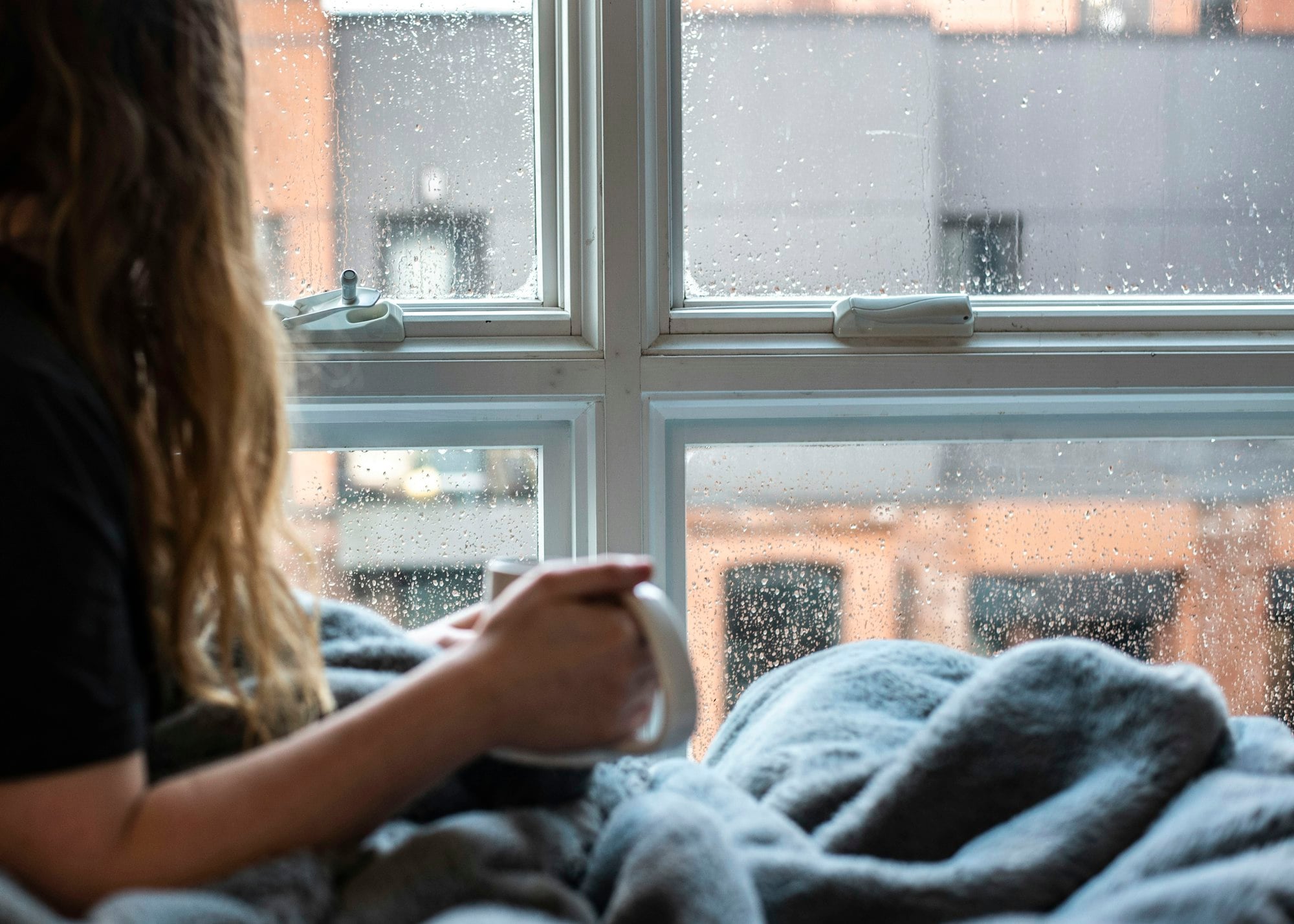A woman with a warm mug looking out a rainy window, pausing to notice what is going on inside
